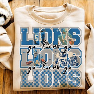 May include: A cream-colored sweatshirt featuring the word "LIONS" in blue and gray block letters, with the phrase "go lions go" in a script font. The design includes images of football players.