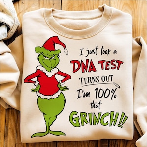 May include: A beige sweatshirt featuring a green Grinch character in a red Santa suit and hat. Text reads "I just took a DNA test, turns out I'm 100% that Grinch!!" in red and green.