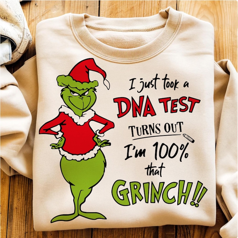May include: A beige sweatshirt featuring a green Grinch character in a red Santa suit and hat. Text reads "I just took a DNA test, turns out I'm 100% that Grinch!!" in red and green.