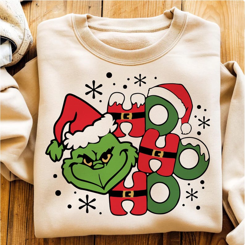 May include: A beige sweatshirt featuring a Grinch graphic wearing a Santa hat and the words "HO HO HO" in red and green. Black snowflakes and dots are scattered around the design. The sweatshirt is folded on a wooden surface.