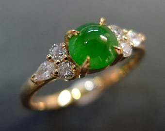 May include: A gold ring featuring a large, vibrant green gemstone at the center, surrounded by smaller, clear gemstones. The ring's band is a warm gold color, and the gemstones sparkle against a neutral background. A piece of jewelry.