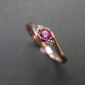 May include: A delicate rose gold ring with a pink ruby gemstone and two smaller diamonds. The ring has a curved band.