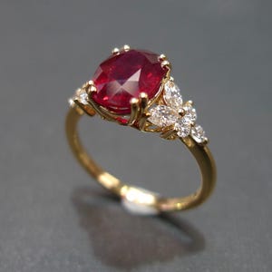 Puede incluir: Un anillo de oro con una gran piedra de rubí ovalada rodeada de diamantes más pequeños. El anillo tiene un diseño floral delicado.