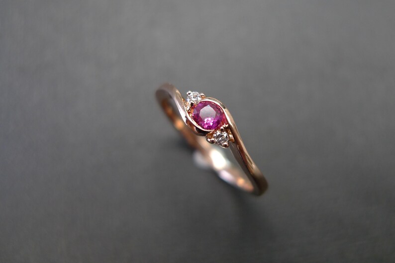 May include: A delicate rose gold ring with a pink ruby gemstone and two smaller diamonds. The ring has a curved band.