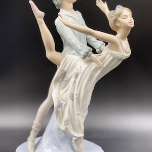 Puede incluir: Figura de porcelana que representa a una pareja de ballet en una pose elegante. La mujer lleva un vestido blanco con detalles florales, y el hombre una camisa azul claro. La base es un diseño de ola azul claro. La figura mide aproximadamente 30 cm de altura.