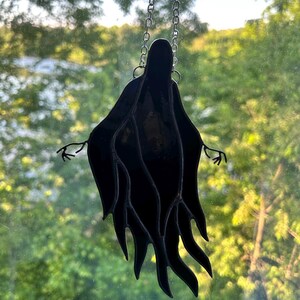 Pu&ograve; includere: Decorazione a fantasma in vetro colorato nero appesa a una catena d'argento. Il fantasma ha le braccia tese e un design a forma di veste fluente. Lo sfondo &egrave; una vista sfocata di alberi verdi e uno specchio d'acqua.