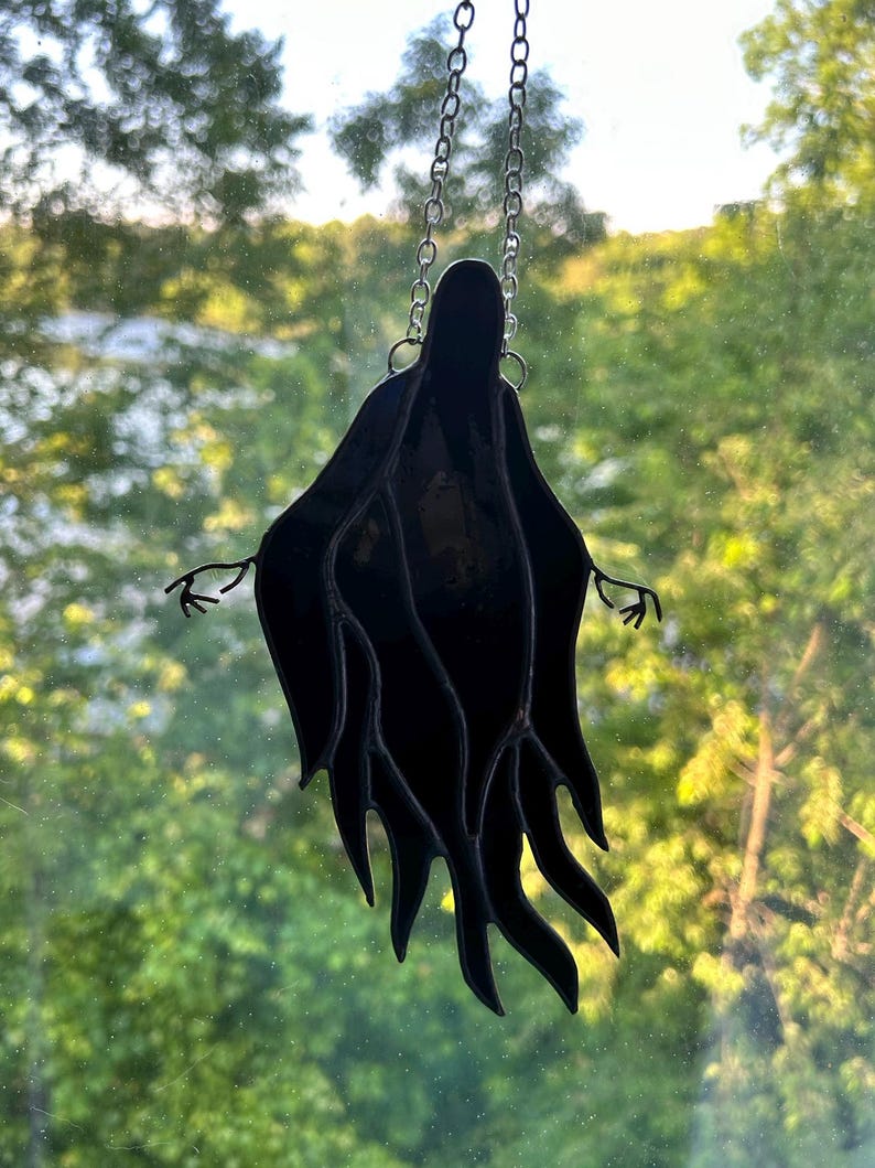 Pu&ograve; includere: Decorazione a fantasma in vetro colorato nero appesa a una catena d'argento. Il fantasma ha le braccia tese e un design a forma di veste fluente. Lo sfondo &egrave; una vista sfocata di alberi verdi e uno specchio d'acqua.
