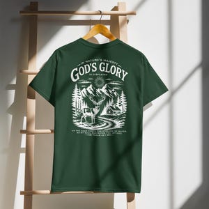May include: Dark green t-shirt with a white graphic of a deer, mountains, trees, and a house. The text on the shirt reads "IN NATURE'S MAJESTY - GOD'S GLORY IS DISPLAYED" and a Bible verse. The t-shirt is hanging on a wooden hanger.