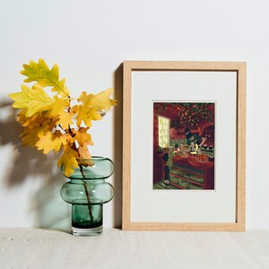 May include: A framed print with a light wood frame depicts a kitchen scene with people. A green glass vase holds yellow and orange leaves. The vase has a rounded, stacked design. The background is a white wall.