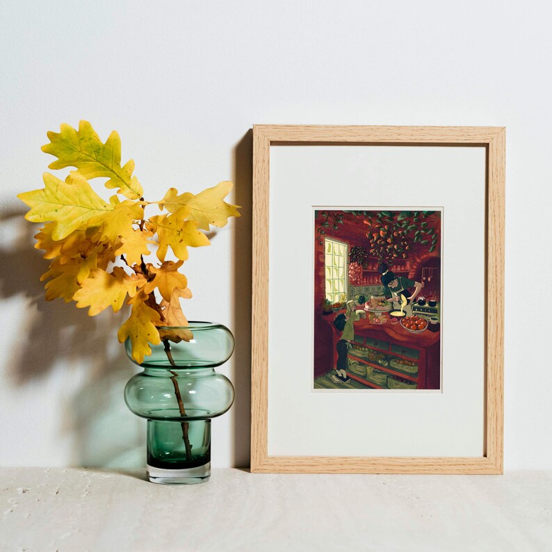 May include: A framed print with a light wood frame depicts a kitchen scene with people. A green glass vase holds yellow and orange leaves. The vase has a rounded, stacked design. The background is a white wall.