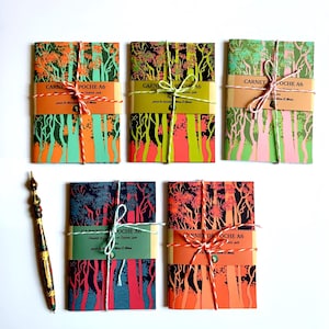 Op de afbeelding: Vijf A6-zaknotitieboekjes met bosillustraties in diverse kleurenpaletten, elk omwikkeld met een papieren band met het label CARNET DE POCHE A6 en vastgebonden met touw, vergezeld van een decoratieve pen met een patroon in meerdere kleuren.