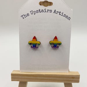 May include: A pair of wooden stud earrings in the shape of a fleur-de-lis, painted with rainbow colors. The earrings are displayed on a white card with the text "The Upstairs Artisan". The card is propped up on a small wooden easel.
