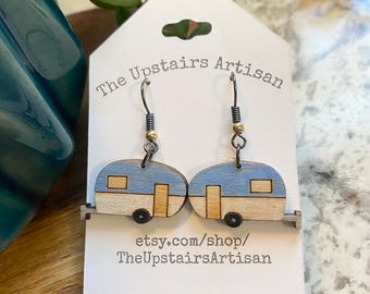 May include: A pair of dangle earrings featuring wooden camper trailer charms painted in light blue and natural wood tones. The earrings hang from dark metal hooks on a white display card printed with The Upstairs Artisan and etsy.com/shop/TheUpstairsArtisan.