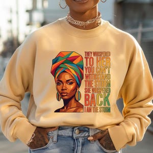 May include: A beige sweatshirt featuring a graphic of a woman wearing a colorful head wrap and earrings. Text on the right reads "THEY WHISPERED TO HER YOU CAN'T WITHSTAND THE STORM SHE WHISPERED BACK I AM THE STORM."
