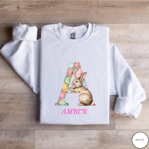 May include: White sweatshirt featuring a colorful patchwork letter "A" with a brown bunny illustration. The name "AMBER" is printed in pink below the bunny. The sweatshirt is on a wooden surface.