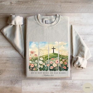 Puede incluir: Sudadera de color arena con un diseño de acuarela que presenta una cruz sobre una colina, rodeada de flores. El texto "HE IS NOT HERE. HE HAS RISEN. Matthew 28:6" está impreso debajo. Las mangas están enrolladas.