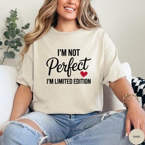 May include: Sand-colored t-shirt with the text "I'M NOT PERFECT I'M LIMITED EDITION" in black script and a red heart. The shirt is worn with blue jeans. The overall theme is casual wear.