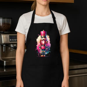 May include: Black apron featuring a colorful illustration of a toy nutcracker soldier. The nutcracker is depicted in pink, blue, and gold, set against a vibrant watercolor splash. The apron has a bib and waist ties, suitable for kitchen or craft use.