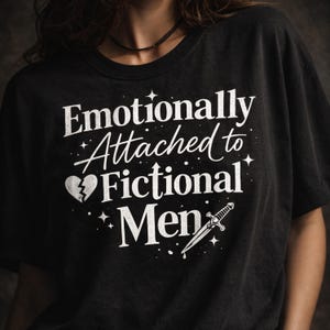 以下が含まれることがあります： 「Emotionally Attached to Fictional Men」の白文字がプリントされた黒いTシャツ。デザインには、割れたハート、星、短剣が含まれています。