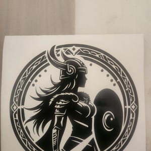 Peut inclure: Autocollant noir et blanc représentant une silhouette de femme guerrière dans un cadre circulaire. La guerrière est de profil, portant un casque à cornes et tenant un bouclier et une épée. Le design présente des détails complexes et une bordure décorative.