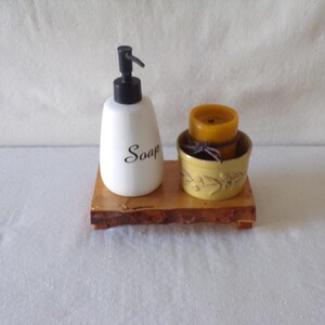 May include: A white soap dispenser with a black pump and the word "Soap" in black script sits on a wooden tray. A yellow candle and a small yellow bowl with a decorative design are also on the tray.
