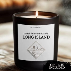 May include: A black glass candle with a white label featuring the text VITEO CANDLE, FOR MOMENTS WHEN YOU MISS LONG ISLAND, and a line art illustration of a lighthouse, sun, and ocean inside a diamond shape. The candle has a lit wick with smoke.