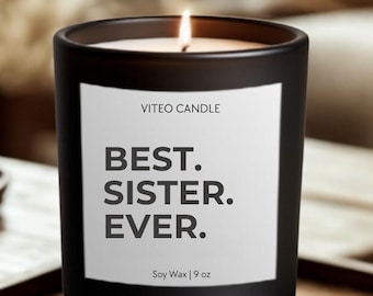 May include: A matte black candle in a glass jar with a white label featuring the text VITEO CANDLE, BEST. SISTER. EVER., and Soy Wax | 9 oz. The candle has a single lit wick, and the wax is white. The container is cylindrical with a smooth finish.