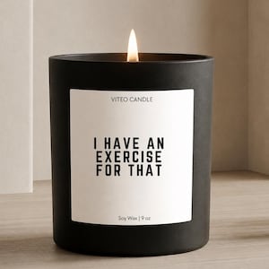 May include: A matte black candle in a cylindrical container with a white label featuring the text VITEO CANDLE, I HAVE AN EXERCISE FOR THAT, and Soy Wax | 9 oz. A single flame is lit at the top of the candle, emitting a warm glow.