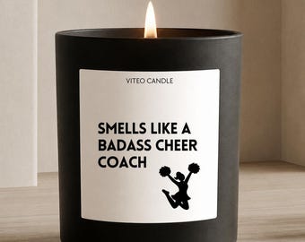 May include: A matte black candle in a cylindrical glass container with a white label. The label features the text VITEO CANDLE, SMELLS LIKE A BADASS CHEER COACH, and a black silhouette of a cheerleader jumping with pom-poms. A flame is lit.