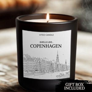 May include: A black matte glass candle with a white label featuring the text VITEO CANDLE, SMELLS LIKE... COPENHAGEN, and a black and white line drawing of a harbor with boats and buildings. The candle is lit, and a black graphic says GIFT BOX INCLUDED.