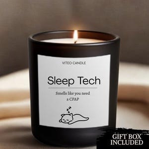 May include: A matte black candle in a glass jar with a white label. The label reads VITEO CANDLE, Sleep Tech, Smells like you need a CPAP, and features a line drawing of a sleeping cat. The candle has a lit wick and a white wax surface.