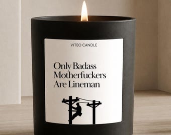 May include: A matte black candle in a cylindrical container with a white label. The label features the text VITEO CANDLE, the phrase Only Badass Motherfuckers Are Lineman, and a black silhouette graphic of a person working on a utility pole.