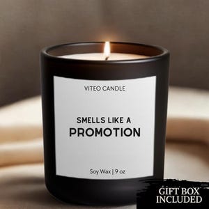 May include: A matte black candle in a glass jar with a white label reading VITEO CANDLE, SMELLS LIKE A PROMOTION, and Soy Wax | 9 oz. The candle has a lit wick with a visible flame. A black graphic in the corner states GIFT BOX INCLUDED.