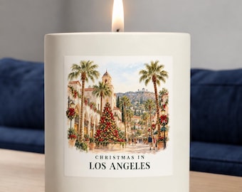 May include: A white cylindrical candle with a lit wick featuring a square label depicting a Christmas scene in Los Angeles with palm trees, a decorated tree, and the text CHRISTMAS IN LOS ANGELES. The candle is placed on a wooden surface.