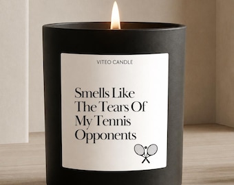 Puede incluir: Una vela cilíndrica de color negro mate con una etiqueta rectangular blanca. La etiqueta presenta el texto VITEO CANDLE, Smells Like The Tears Of My Tennis Opponents, y un pequeño gráfico de dos raquetas de tenis cruzadas con una pelota. Una llama está encendida.