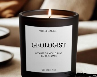 May include: A matte black candle in a glass jar with a white label reading VITEO CANDLE, GEOLOGIST, BECAUSE THE WORLD RUNS ON ROCK STARS, and Soy Wax | 9 oz. The candle has a lit wick and is presented with a gift box included notice.