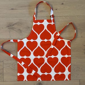 以下が含まれることがあります： 赤と白のハート模様のエプロン。エプロンには首ひもとウエストタイが付いています。ハート模様は、白い背景に赤いハートで構成されています。エプロンは布製で、キッチンでの使用を目的としています。