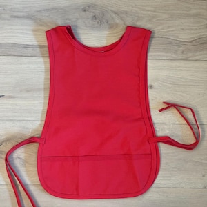 May include: A solid red apron with a rounded bottom edge and a small pocket. The apron has a scoop neckline and ties at the waist. The apron is made of a smooth, solid fabric.