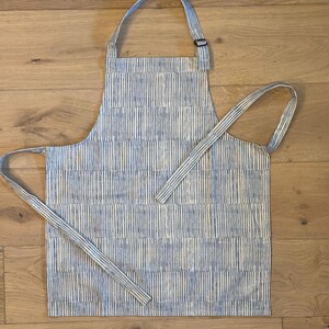 以下が含まれることがあります： 青と白のストライプのエプロン。調節可能なネックストラップとウエストタイが付いています。エプロンは、垂直の青いストライプが入った明るい色の生地で作られています。キッチンでの使用向けに設計されています。
