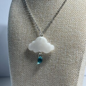 May include: A silver chain necklace with a white cloud pendant and a blue teardrop charm. The cloud pendant is a puffy, irregular shape. The teardrop charm is a translucent blue color.