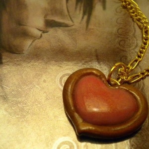 May include: A gold chain necklace with a heart-shaped pendant. The pendant is made of brown clay and has a pink heart inside.