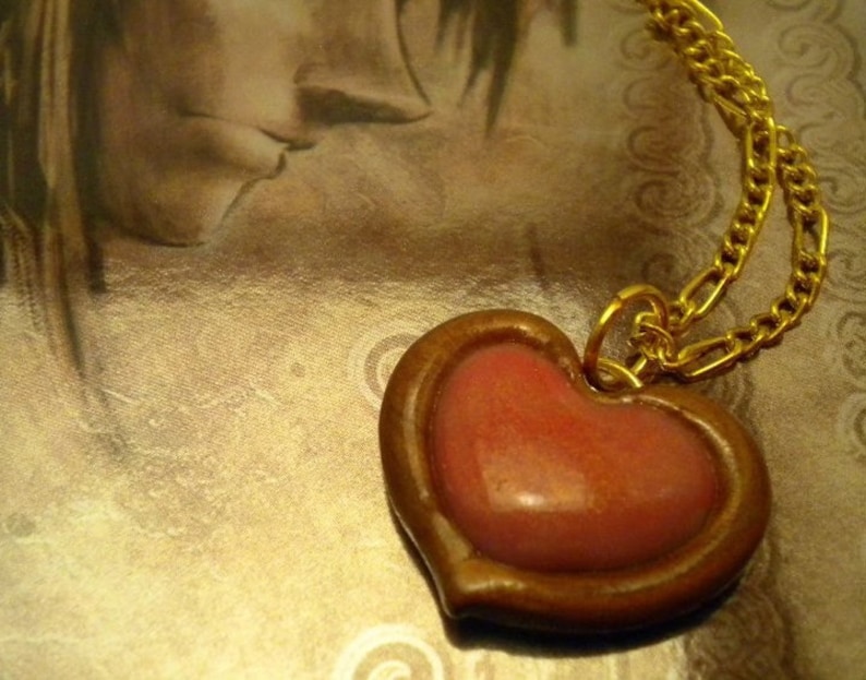 May include: A gold chain necklace with a heart-shaped pendant. The pendant is made of brown clay and has a pink heart inside.
