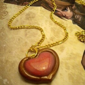 May include: A gold chain necklace with a heart-shaped pendant. The pendant is made of brown clay with a pink and gold glitter finish.