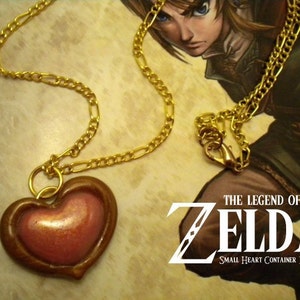 May include: A gold chain necklace with a heart-shaped pendant. The pendant is made of brown clay with a pink heart inside. The text "THE LEGEND OF ZELDA SMALL HEART CONTAINER NECKLACE" is visible in the background.