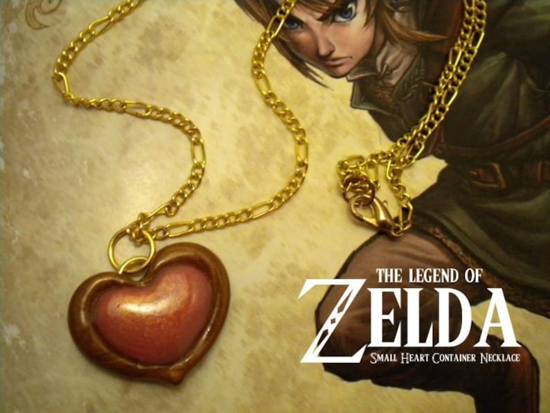 May include: A gold chain necklace with a heart-shaped pendant. The pendant is made of brown clay with a pink heart inside. The text "THE LEGEND OF ZELDA SMALL HEART CONTAINER NECKLACE" is visible in the background.
