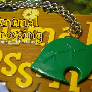 May include: A green leaf pendant necklace with a silver chain. The pendant is shaped like the iconic leaf from the Animal Crossing video game series. The text "Animal Crossing Tom Nook Leaf Necklace" is visible in the background.