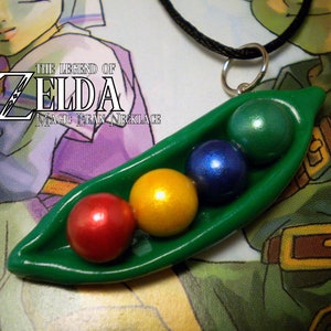 May include: A green pea pod necklace with five colorful beads inside. The beads are red, yellow, blue, and green. The necklace is made of black cord and has a silver clasp. The necklace is labeled "The Legend of Zelda Magic Bean Necklace".