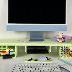 以下が含まれることがあります： 明るい緑色のコンピューターモニター台。収納コンパートメントが内蔵されています。モニター、キーボード、マウス、文房具などを置くことができます。スマートフォンには通知が表示されています。小さなウサギの置物が上に置かれています。