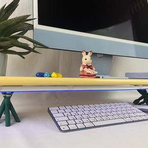 以下が含まれることがあります： ライトブルーのコンピューターモニター、青色LEDストリップ付きの黄色い棚、白いキーボードを備えたモダンなデスクセットアップ。小さなウサギの置物と鉢植えの多肉植物が装飾的なタッチを追加します。デスクは緑色の三脚脚で支えられています。