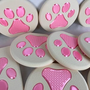 May include: A collection of off-white, paw-shaped coasters with pink paw prints. The paw pads have a textured design. The coasters are round and oval, and are arranged in a close-up shot.